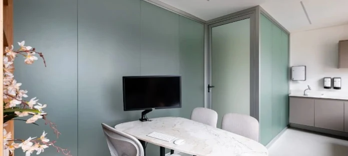 Outra vista do consultório, com mesa clara, cadeiras e painéis em tom verde suave.